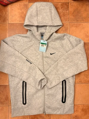 Nike x NOCTA Tech Fleece Hoodie - Heather Grey - Helt ny! - Säljer en helt ny och oanvänd Nike x NOCTA Tech Fleece hoodie i den klassiska färgen Heather Grey. Detta är ett exklusivt samarbete mellan Nike och Drake (NOCTA) som kombinerar teknisk funktion med en stilren, sportig estetik. Tröjan har en unik passform och detaljer som skiljer den från den vanliga Tech Fleece-linjen. 