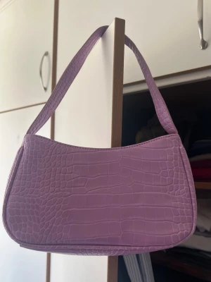 Ljuslila axelväska i krokopräglat skinn - Trendig ljuslila handväska med axelrem och croco-mönstrad yta. Väskan har en stilren, rektangulär form och är tillverkad i syntetmaterial som liknar skinn. Perfekt accessoar för att lyfta din outfit.