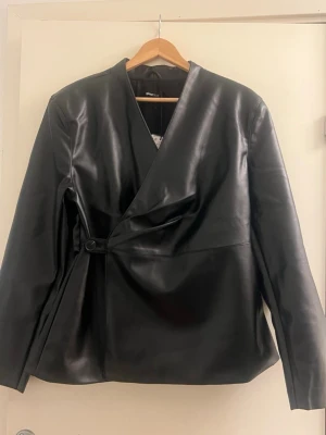 Svart omlott skinnjacka från Gina Tricot - Snygg svart skinnjacka från Gina Tricot  med omlottknäppning och minimalistisk design. Jackan har en enkel knappdetalj i sidan, rak passform och mjukt, glansigt skinnmaterial. Perfekt för dig som gillar stilrena och moderna plagg.