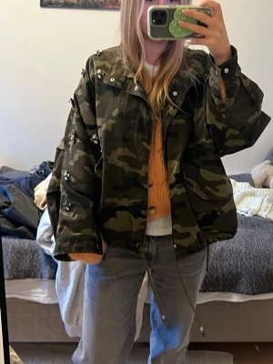 Jeansjacka med militärmönster och nitar - Snygg oversize jacka i camomönster med gröna och bruna toner. Jackan har stora fickor, dragkedja och knappar framtill samt coola pärlor och nitar på axlar och ärmar. Materialet är kraftigt tyg som påminner om denim men är mjukare. Perfekt för dig som vill ha en unik streetstyle.