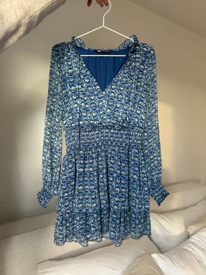 Blå mönstrad kortklänning med volang - Säljer en blå kortklänning från Zara med vitt och grönt blommigt mönster. Klänningen har v-ringning, volangdetaljer vid halsen och ärmslut samt långa ärmar. Den är tillverkad i ett lätt och luftigt material, perfekt för vår och sommar.