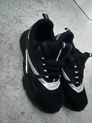 Svarta sneakers med silverdetaljer - Snygga svarta sneakers med coola silverdetaljer och mesh på ovansidan. Skorna har tjock sula och klassiska svarta skosnören. Perfekt för dig som vill ha en modern och sportig look.