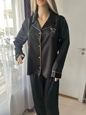 Svart satinpyjamas med rosa detaljer - Svart pyjamas i satin från Victoria's Secret med rosa kantband och knappar. Setet har långärmad skjorta med klassisk krage och VS-logga i strass på bröstet samt matchande byxor. Perfekt för mysiga kvällar och stilren lounging.