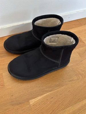 Svarta tofflor från McKinley - Svarta tofflor från McKinley med mjukt och varmt foder i beige. Tofflorna har en klassisk rundad form och är tillverkade i mockaliknande material med robust sula. Perfekta för att hålla fötterna varma under kalla dagar.