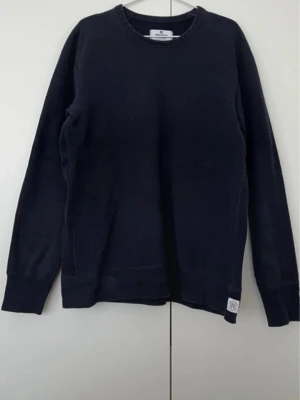 Svart stickad tröja från Reigning Champ - En stilren svart stickad tröja från Reigning Champ med rund hals och ribbade muddar. Tröjan har en diskret vit patch med logotypen på nederkanten. Perfekt för dig som gillar enkel och klassisk design.