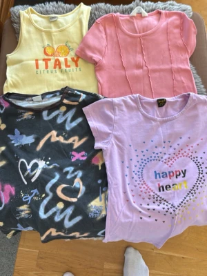 Fyra coola t-shirts i olika färger - Säljer fyra t-shirts: en gul ärmlös med citrusmotiv och texten 'ITALY', en rosa kortärmad med vågiga sömmar, en svart kortärmad med färgglada abstrakta mönster och en ljuslila kortärmad med hjärttryck och texten 'happy heart'. Perfekt för dig som gillar färg och mönster!