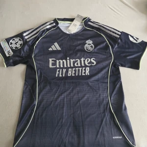 Real Madrid matchtröja Mbappé #10 - Säljer en mörkblå Real Madrid matchtröja från Adidas med Mbappé och nummer 10 på ryggen. Tröjan har vita detaljer, klubbmärke och sponsortryck på framsidan samt patchar på ärmarna. Materialet är lätt och andas, perfekt för fotboll eller träning.