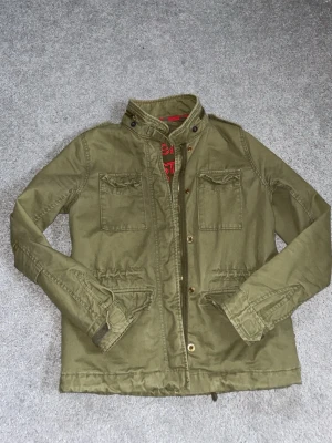 Olivgrön overshirt från Superdry - Stilren olivgrön field jacket från Superdry med broderad logga och text på insidan. Två bröstfickor med lock, samt brittiska flaggan på axeln. Passar perfekt på sommarkvällar eller vår!