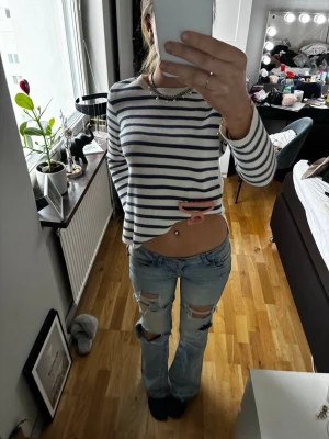 Randig långärmad tröja i vitt och blått - Säljer en snygg långärmad tröja med vita och mörkblå horisontella ränder. Tröjan har en rak passform och rund halsringning, perfekt för en chill och stilren look. Materialet är ribbstickat och mjukt, vilket gör den bekväm att bära.