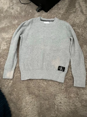 Ljusgrå stickad tröja Calvin Klein - Snygg ljusgrå stickad tröja från Calvin Klein med rund halsringning och ribbade muddar. Tröjan har en svart patch med CK-logga nedtill på framsidan. Perfekt för dig som gillar enkel och stilren design.