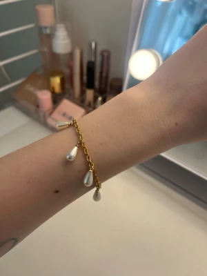 Guldigt armband med pärldroppar - Snyggt armband i guldfärgad metall med fem hängande droppformade pärlor. Kedjan är tunn och har ett klassiskt lås. Perfekt för att ge din look en elegant touch. Passar dig som gillar stilrena accessoarer med lite extra detalj. Handgjort och rostfritt stål