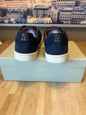 Mörkblå sneakers från Brunello Cucinelli - Stilrena mörkblå sneakers i mocka från Brunello Cucinelli med vit sula och guldfärgad logga broderad på hälen. Insidan har bruna detaljer och skorna har en klassisk låg profil som passar till många olika outfits.