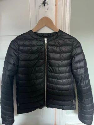 Svart jacka - Snygg svart pufferjacka med quiltad design och raka sömmar. Jackan har dragkedja framtill och två sidofickor med dragkedja. Tillverkad i ett glansigt, lätt material som håller värmen utan att kännas tung. Perfekt för kalla vinterdagar.