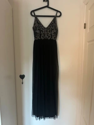 Svart långklänning med pärldetaljer - Elegant svart långklänning med smala axelband och djup V-ringning. Överdelen är dekorerad med glittrande pärlor i ett detaljerat mönster och kjolen är i tunt, flytande tyg. Perfekt för bal eller festkväll.