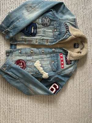 Superdry ljusblå jeansjacka xxs - Cool ljusblå jeansjacka från Superdry med massor av broderade patches och detaljer, bland annat siffror, stjärnor och brittiska flaggan. Jackan har fuskpäls på insidan av kragen och stora Superdry-bokstäver på ryggen. Perfekt för dig som gillar streetstyle och unika plagg. Nypris 899kr säljs inte längre. 