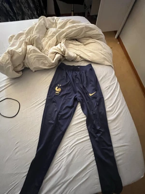 Blå Nike träningsbyxor Frankrike - Säljer ett par blå träningsbyxor från Nike med Frankrikes landslagslogga på ena sidan och Nike-loggan i guld på andra. Byxorna är smala i modellen och har elastisk midja, perfekta för fotboll eller annan sport.