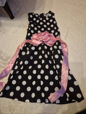 Barn kläder  - En svart kortklänning med vita prickar och ett brett rosa sidenband med stor rosett i midjan. Klänningen är ärmlös och har en klassisk, lekfull design som passar perfekt för dig som gillar retrostil och att sticka ut. I olika storlekar 