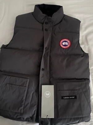 Svart dunväst från Canada Goose - Snygg svart dunväst från Canada Goose med hög krage, två stora fickor framtill och klassisk logopatch på bröstet. Västen har tryckknappar och är tillverkad i slitstarkt material med dunfyllning som håller dig varm. Perfekt för lager på lager och streetwear-vibe.