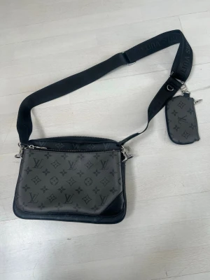 Monogram axelväska svart - Snygg axelväska i svart och grått med klassiskt monogrammönster. Väskan har justerbar bred axelrem och en liten avtagbar ficka. Tillverkad i canvas med läderdetaljer och silverfärgade metalldetaljer.