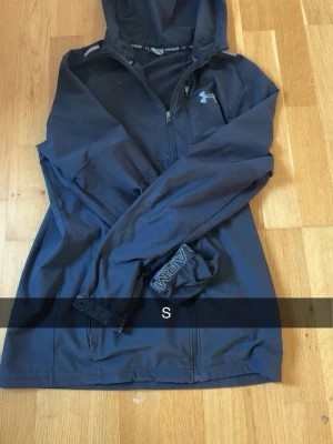 Svart Under Armour hoodie med dragkedja - Svart hoodie från Under Armour med dragkedja och huva. Logotypen syns på bröstet och ärmen har tryck med 'ARMOUR'. Perfekt för träning eller chill, tillverkad i ett lätt och stretchigt material som ger bra rörelsefrihet.