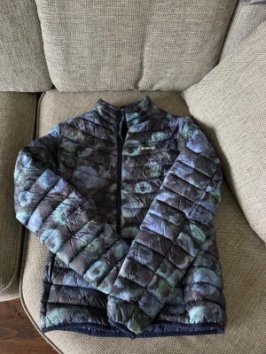 Mönstrad dunjacka från Everest - Snygg dunjacka från Everest med unikt mönster i blå, lila och gröna toner. Jackan har hög krage, dragkedja framtill och lätt vadderad design som håller dig varm under kalla dagar. Perfekt för dig som vill sticka ut i vinterkylan.