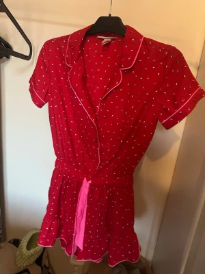 Röd kortärmad pyjamas med mönster - Söt röd pyjamas med korta ärmar och ben, små vita mönster och vita passpoaler längs kanterna. Har en rosa midjerem för extra detalj och en avslappnad krage. Perfekt för mysiga kvällar hemma.