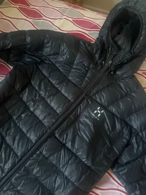 Svart dunjacka från Haglöfs med huva - Snygg svart dunjacka från Haglöfs med quiltad design och fast huva. Jackan har dragkedja framtill och en bröstficka med dragkedja. Perfekt för kalla vinterdagar och riktigt skön att bära. Klassisk logga på bröstet. Kan gå ner i pris vid snabbbäffär kan mötas upp Helsingborg/malmö annars tillkommer frakt pris 