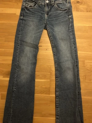 Blå bootcut jeans med slitningar - Säljer ett par blå bootcut jeans med snygga slitningar och klassiska femficksdetaljer. Jeansen har en normal passform och är tillverkade i denim med ljus tvätt. Perfekta för dig som gillar en avslappnad och tidlös look.