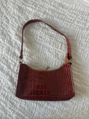 Brun axelväska i krokopräglat skinn - Snygg brun handväska med axelrem i krokopräglat konstläder. Väskan har en stilren, rektangulär form och stängs med dragkedja i guld. Perfekt accessoar för att lyfta din outfit.
