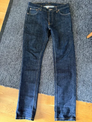 Mörkblå jeans från Nudie Jeans - Snygga mörkblå jeans från Nudie Jeans med klassisk femficksmodell och raka ben. Jeansen har kontrastsömmar och ikoniska broderade bakfickor. Tillverkade i slitstark denim, perfekta för en trendig och avslappnad stil. Två mindre defekter se bild 