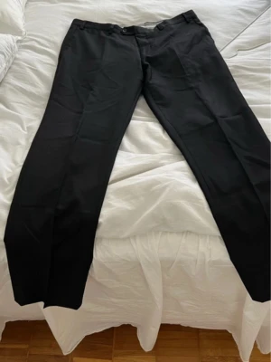 Svarta kostymbyxor från The Super Outfit - Snygga svarta kostymbyxor från The Super Outfit med klassisk rak passform och diskreta pressveck. Perfekta för dig som vill ha en stilren look. Materialet är mjukt och bekvämt, vilket gör dem lätta att bära hela dagen.