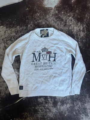 Vit tröja från Morris med tryck - Vit långärmad tröja från Morris med tryck framtill som inkluderar brittisk flagga och text. Tröjan har rund halsringning, ribbade muddar och är tillverkad i ett stickat material. Perfekt för en stilren och klassisk look.