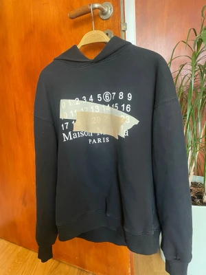 Maison Margiela svart hoodie - Svart hoodie från Maison Margiela med ikoniskt tryck av siffror och beige tejpdetalj på bröstet. Oversized passform, ribbade muddar och huva. Perfekt för dig som gillar streetwear och unika designer. Materialet är mjukt och bekvämt bomull.