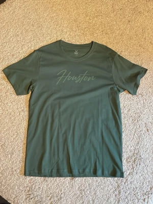 Grön t-shirt från SOC med tryck - Snygg grön t-shirt från SOC med diskret 'Houston'-tryck på bröstet. Klassisk rund hals och korta ärmar, perfekt för dig som gillar enkel och stilren design. Materialet är mjukt och känns skönt mot huden.