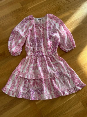  Ny slutsåld Rosa och vit kortklänning med volang - Söt kortklänning med rosa och vitt paisleymönster, volangdetaljer och v-ringad hals. Klänningen har trekvartsärmar och knytband i midjan för en snygg passform. Perfekt för sommarens alla tillfällen!