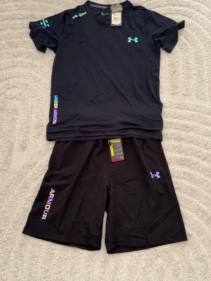 Svart träningsset från Under Armour - Snyggt svart träningsset från Under Armour med t-shirt och shorts. Setet har färgglada loggor och textdetaljer i regnbågsfärger. Tillverkat i ett lätt och snabbtorkande funktionsmaterial som passar perfekt till träning och sport.