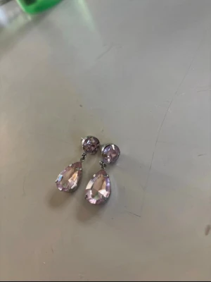 Ljuslila droppformade örhängen - Säljer ett par glittrande örhängen med ljusrosa stenar. Örhängena har en rund sten upptill och en större droppformad sten som hänger ned. Perfekta för dig som gillar att sticka ut med lite extra bling. Metallfärgad infattning.