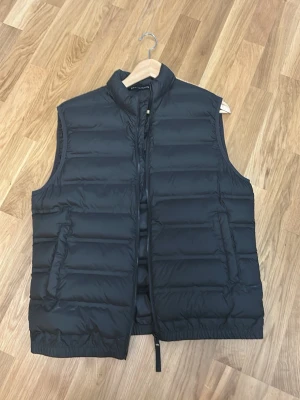 Svart dunväst från Zara - Säljer en svart dunväst från Zara med quiltad design och hög krage. Västen har två sidofickor och dragkedja framtill. Perfekt för lager-på-lager och håller dig varm under kyliga dagar. Materialet är lätt och smidigt, med klassisk passform.