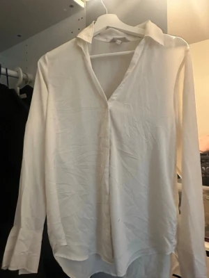 Vit långärmad blus med krage - Stilren vit blus med klassisk krage och v-ringning. Blusen har lång ärm och lätt rundad nederkant. Materialet ser ut att vara tunt och mjukt, perfekt för en clean och enkel look.