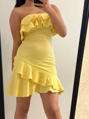 Gul axelbandslös volangklänning - Snygg gul kortklänning med volanger upptill och längs kjolen. Axelbandslös design som ger en fräsch och somrig look. Klänningen har en figurnära passform och är tillverkad i ett mjukt, stretchigt material som sitter bekvämt.