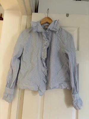 Blårandig blus med volangkrage - Snygg blårandig blus från H&M med volangdetaljer längs knäppningen och en hög volangkrage. Blusen har långa ärmar med breda manschetter