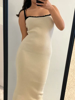 Ribbad beige midiklänning - Trendig ribbad midiklänning i beige med smala axelband och svart kantdetalj vid urringningen. Figurnära passform och stretchigt material som ger en snygg siluett. Perfekt för dig som gillar minimalistisk och stilren look.