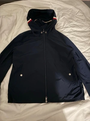 Mörkblå vindjacka från Moncler - Snygg mörkblå vindjacka från Moncler med huva och dragkedja framtill. Jackan har två fickor med tryckknappar och Moncler-logga på bröstet. Detaljer i rött och vitt på huvan ger en sportig touch. Perfekt för blåsiga dagar.
