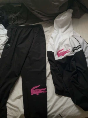 Svart och vit tracksuit från Lacoste - Säljer en snygg tracksuit från Lacoste med svart byxa och svartvit jacka med huva. Båda delarna har stora rosa Lacoste-loggor och jackan har även en svartvit Lacoste-text på ärmen. Perfekt för träning eller chill.