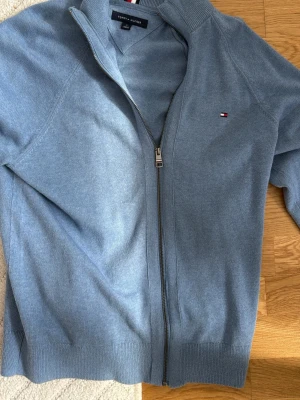Blå ziptröja från Tommy Hilfiger - Snygg ljusblå ziptröja från Tommy Hilfiger med diskret logga på bröstet. Tröjan har ribbade muddar och krage, hel dragkedja och är tillverkad i mjukt stickat material. Perfekt för lager-på-lager och en clean look. NY PRIS ÄR 1300 KÖPT I FRANKRIKE