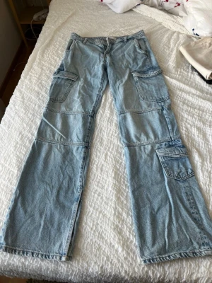 Ljusa denim cargobyxor med fickor från zara - Säljer ett par ljusa cargobyxor i denim med flera stora fickor på benen och coola sömdetaljer. Byxorna har en avslappnad passform och är perfekta för dig som gillar streetstyle och funktionella kläder. Materialet är klassisk jeans och färgen är ljusblå.