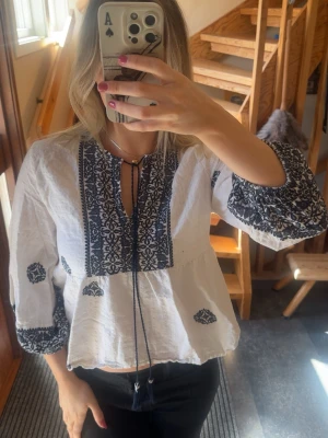 Vit boho-blus med blå brodyr - Vit boho trekvartärmsblus med mörkblå broderade detaljer och knytband vid halsen. Från Zara i storlek M, bra skick förutom att den har en ”prick” (se sista bilden) 🌻