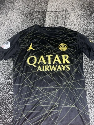Svart PSG matchtröja Jordan - Snygg svart PSG matchtröja med gula detaljer och grafiskt mönster. Tröjan har PSG-logga, Jordan-logga och Qatar Airways tryck på bröstet samt GOAT och Ligue 1-märke på ärmen. Tillverkad i ett lätt och ventilerande material, perfekt för fotboll eller träning.