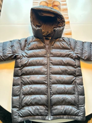 Svart dunjacka från Peak Performance - Snygg svart dunjacka från Peak Performance med huva och dragkedja framtill. Jackan har två sidofickor med dragkedja och ribbade muddar vid ärmsluten. Perfekt för kalla vinterdagar och riktigt skön att bära.