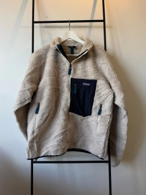 Beige teddyjacka från Patagonia - Mysig beige teddyjacka från Patagonia med halv dragkedja och hög krage. Jackan har en svart bröstficka med dragkedja och två sidofickor med blå detaljer. Perfekt för kalla dagar och har en avslappnad passform.